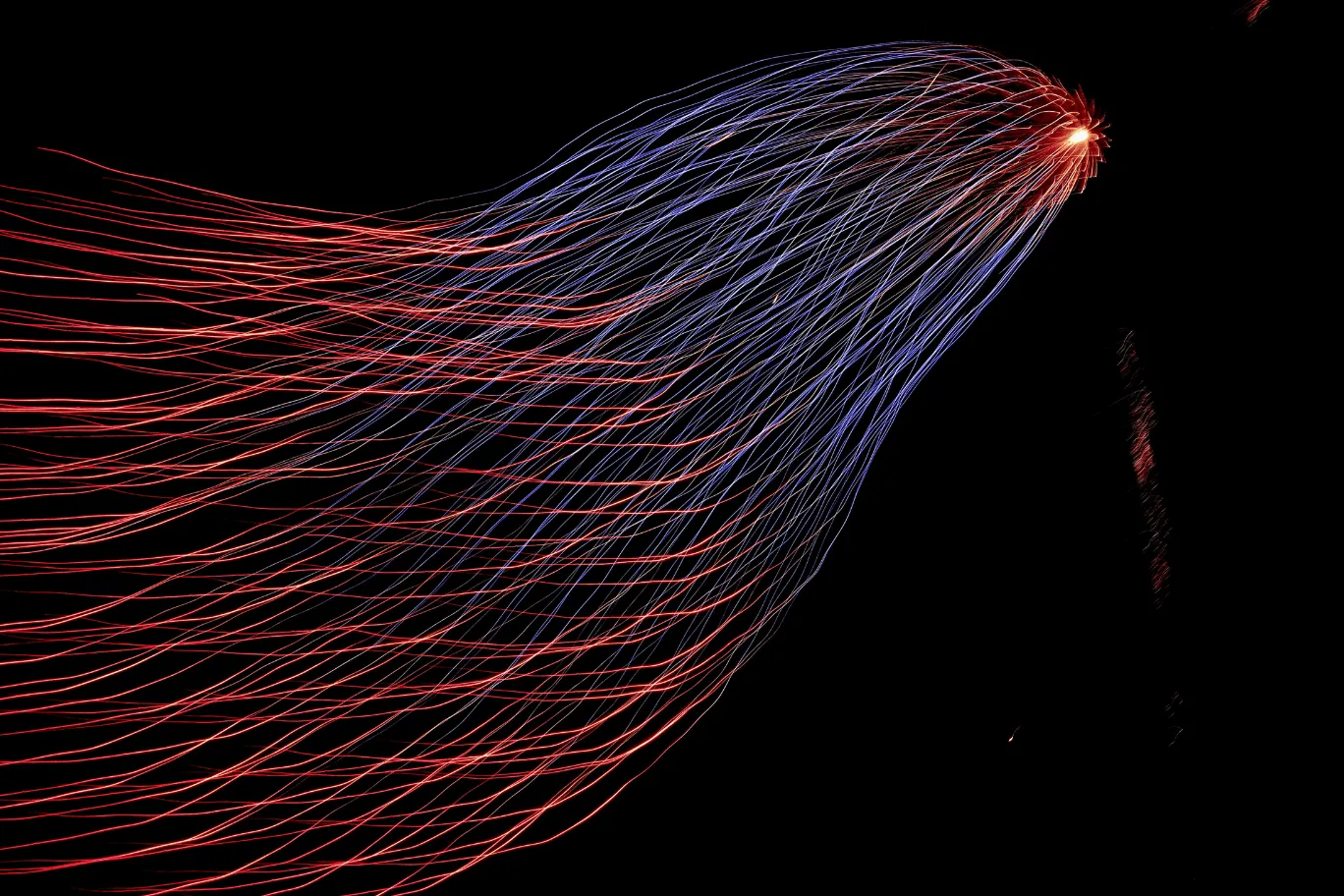 Long-exposure photograph of red and blue sparkler trails fanning outward from a single bright point against a black sky